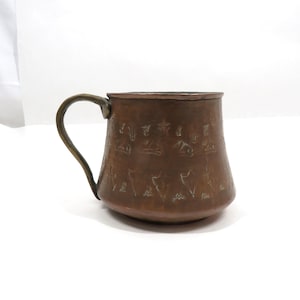 May include: A copper-colored mug with a curved handle. The mug has an embossed design around the body. The handle is a darker shade of copper. The mug is sitting on a white surface.