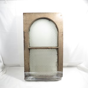 May include: An arched wooden window frame with a weathered finish, featuring two panes of frosted glass with a subtle repeating pattern. The lower section of the frame is wrapped in clear plastic film for protection during transport.