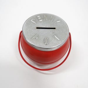 May include: A round red coin bank with a silver-colored metal lid featuring a horizontal coin slot. The lid is embossed with the text PENNY POT in capital letters. A thin red metal handle is attached to the sides of the red container.