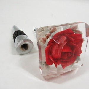 May include: A clear glass bottle stopper with a red rose encased in resin. The stopper has a silver metal top with a threaded screw.