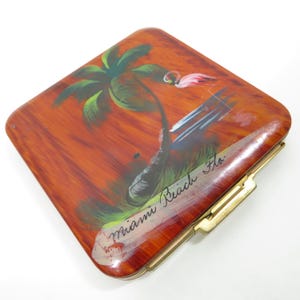 May include: A vintage, square-shaped compact with a painted design of a palm tree and a flamingo. The compact is made of a brown, marbled material and has a gold-coloured clasp. The text "Miami Beach Fla." is painted on the front of the compact.