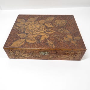 May include: A square, brown wooden box with a floral design carved into the lid. The box features a detailed carving of a rose and leaves, with a brass clasp. The box is a medium brown color.