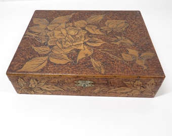 May include: A square, brown wooden box with a floral design carved into the lid. The box features a detailed carving of a rose and leaves, with a brass clasp. The box is a medium brown color.
