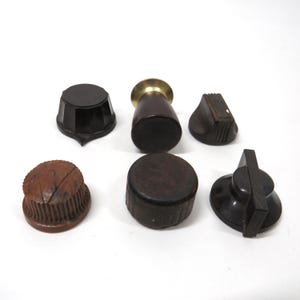 May include: A collection of six vintage radio knobs in various shapes and colors. The knobs are primarily brown and black, with some featuring brass accents. They are arranged on a white surface.