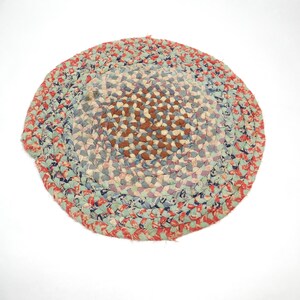 May include: Round braided rug with a mix of colors including red, blue, and brown. The rug is made of fabric strips woven together in a circular pattern. The rug is a decorative item.