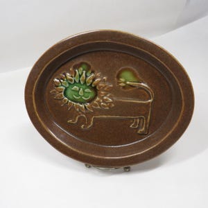 May include: Brown ceramic oval dish with a raised lion design. The lion has a green face and mane, with a long tail. The dish is a decorative piece, likely for display or serving.