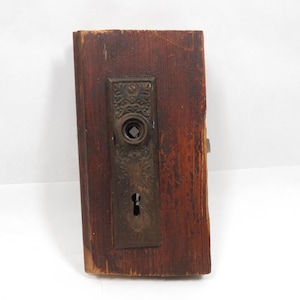 May include: Antique door hardware set on a weathered wooden plank. The set includes a decorative metal plate with a keyhole and a circular knob. The wood has a rich, dark brown color with visible grain and signs of age.