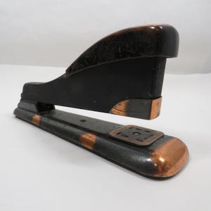 May include: Vintage black and copper stapler. The stapler has a black body with copper accents on the edges and base. The stapler is made of metal and has a classic design. The stapler is a desk accessory.