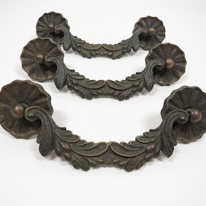 May include: Three antique bronze cabinet pulls with ornate floral designs. Each pull features a round, decorative knob with a raised center and a curved, leaf-shaped handle.