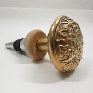May include: A gold bottle stopper with an ornate design. The stopper has a wooden base and a metal top.