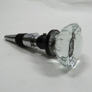 May include: A silver and black bottle stopper with a clear glass knob. The knob has a faceted design.
