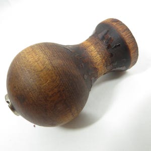 May include: A wooden bottle stopper with a rounded body and a flared top. The wood has a dark, stained finish with visible grain and some areas of peeling. A small metal piece is attached to the base.