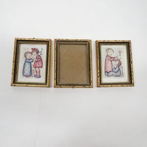 May include: Three vintage gold-framed pictures. Two frames contain illustrations of children in soft colors, one with a cross. The third frame is empty. The frames have a decorative, textured border.