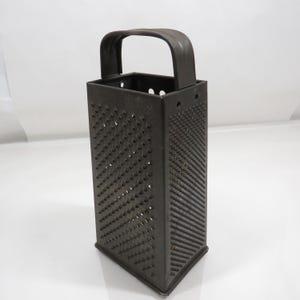 May include: A dark brown, four-sided box grater with a sturdy arched handle at the top. Each side features a uniform pattern of small, sharp, raised perforations for grating food. The metal construction has a matte finish with visible wear.