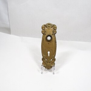 May include: An antique brass door plate featuring an ornate, scrolled design at the top and bottom. The rectangular plate includes a circular opening for a door knob and a vertical keyhole slot below it, with small screw holes for mounting.