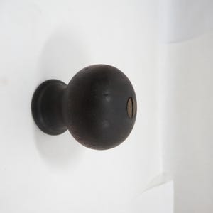 May include: A dark brown, spherical door knob with a circular base. The knob features a small, oval keyhole. The door knob is mounted on a white surface.