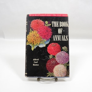 May include: A vintage book titled "The Book of Annuals" by Alfred Carl Hottes. The cover is black with colorful illustrations of various flowers. The book is displayed on a metal stand.