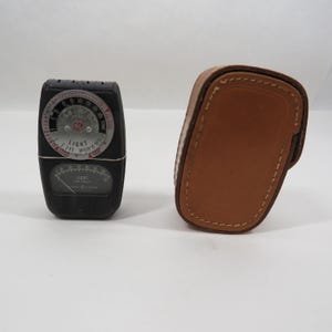 May include: A vintage black light meter with a circular dial and a lower gauge, alongside a brown leather case. The light meter has the word "LIGHT" printed on it.