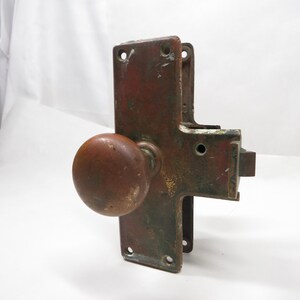 May include: An antique door latch with a round, brown doorknob. The metal components have a weathered, reddish-brown finish with signs of age and use. The latch has a rectangular backplate with mounting holes.