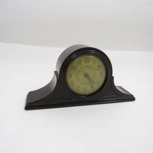 May include: A vintage black mantel clock with a round face and a curved top. The clock face has black numerals and hands on a light green background. The clock is set on a black base.