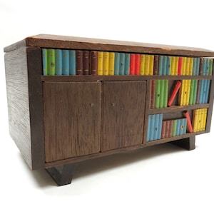 May include: A wooden bookcase with a hinged top and two doors. The bookcase is decorated with colorful painted books. The bookcase is a small decorative box.