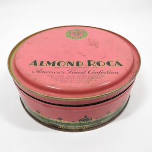 May include: A vintage, round Almond Roca candy tin with a pink lid and sides. The lid features the text "ALMOND ROCA" and "America's Finest Confection" in black lettering. The tin's rim has a green and gold decorative band.