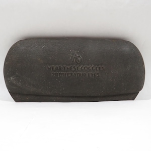 May include: A dark gray, rectangular glasses case with rounded edges. The case has the text "WEAR THESE GOGGLES PROTECT YOUR EYES" printed on the front. The case is made of a textured material.