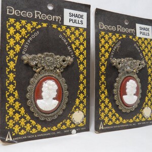 May include: Two vintage shade pulls on their original packaging. Each pull features a cameo design with a white profile against a reddish-brown background, framed by an ornate, aged metal border. The packaging reads "Deco Room" and "SHADE PULLS".