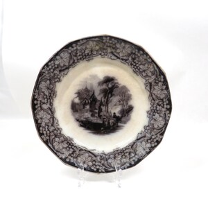 May include: A round ceramic plate with a scalloped edge featuring a black and white transferware pattern. The border displays a detailed grape vine and leaf motif, while the center depicts a pastoral landscape scene with trees and buildings.