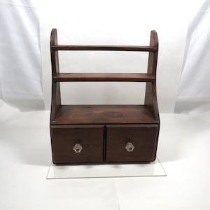 Peut inclure: Une étagère en bois brun foncé avec deux petits tiroirs et deux étagères. Les tiroirs ont des boutons en forme de cristal. L'étagère a une structure en forme de poignée sur le dessus. Le bois a un grain naturel.