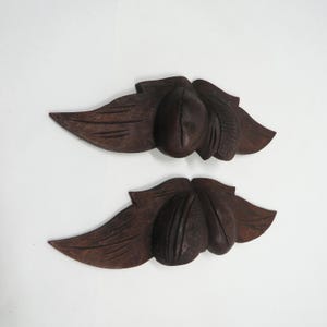 May include: Two dark brown, carved wooden leaf and fruit ornaments. Each ornament features a central fruit shape flanked by two smaller fruit shapes, with a large leaf on each side. The ornaments are set against a white background.