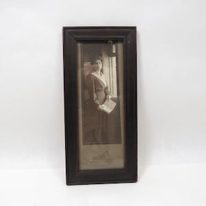 May include: Antique framed black and white photograph of a woman standing by a window, holding a book. The dark wooden frame surrounds the image, which has a sepia tone. The photograph is a vintage piece.