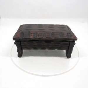 May include: A dark brown wooden box with a textured, carved lid and sides. The box has a rectangular shape and rests on four short legs. The lid features a pattern of rounded indentations, and the wood appears to be aged.