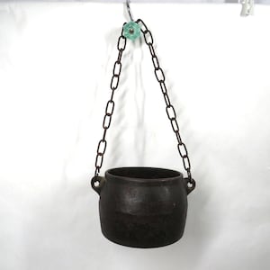 May include: A dark brown, cast iron hanging pot with two handles. The pot is suspended by a rusty chain attached to a light green, decorative knob. The pot is likely used for decorative purposes or for small plants.