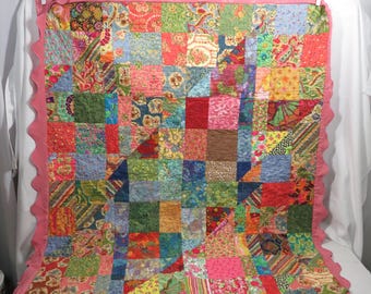 May include: A patchwork quilt with a pink scalloped border. The quilt is made of many different fabrics in various shades of pink, red, green, blue, and brown. The quilt is quilted in a simple pattern.