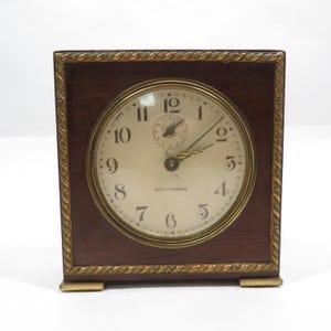 May include: A vintage Seth Thomas clock with a square, dark wood frame and a gold-toned bezel. The clock face is cream-coloured with black numerals and hands. The clock measures approximately 15 cm wide and 15 cm tall.