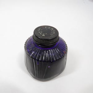 Peut inclure: Un flacon d'encre vintage de couleur violet foncé, avec un design en verre côtelé. Le flacon est surmonté d'un bouchon noir, avec le texte "PARKER" et "INK" visible. L'encre à l'intérieur semble être d'un violet profond et riche.