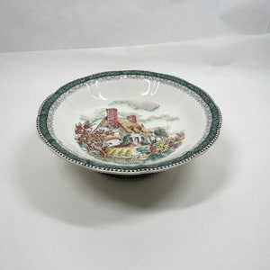 May include: A white ceramic bowl with a green and white border. The bowl features a cottage scene with a red roof and green trees. The bowl has a pedestal base.
