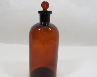 May include: A brown glass bottle with a glass stopper. The bottle is tall and cylindrical with a rounded bottom. The stopper is a small, round knob.
