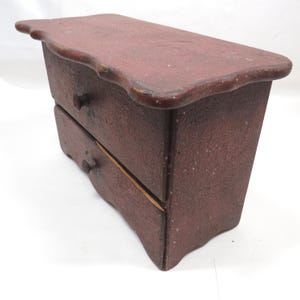 May include: A small, two-drawer wooden chest painted in a deep burgundy color. The chest has a scalloped top and drawer pulls. The wood grain is visible, giving it a rustic appearance. The drawers are visible and appear to be empty.