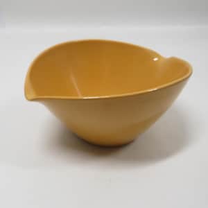 May include: A mustard-yellow ceramic bowl with a unique, asymmetrical shape. The bowl has a smooth, glossy finish and a slightly flared rim. The design features a subtle spout-like extension on one side, adding a touch of elegance to this kitchenware item.