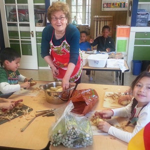May include: A group of children are peeling potatoes in a classroom. A woman in a red apron is supervising them. There is a large pot of potatoes on the table. The children are using a variety of tools to peel the potatoes. There is a German flag in the background.