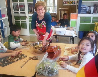 May include: A group of children are peeling potatoes in a classroom. A woman in a red apron is supervising them. There is a large pot of potatoes on the table. The children are using a variety of tools to peel the potatoes. There is a German flag in the background.