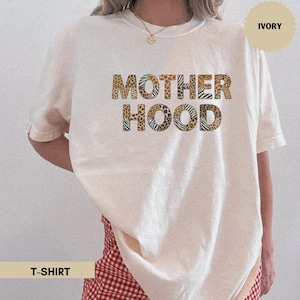 Puede incluir: Camiseta de color marfil con la palabra "MOTHERHOOD" en letras grandes y en negrita. Las letras están rellenas de varios estampados de animales. La camiseta también tiene la palabra "T-SHIRT" en una pancarta en la parte inferior.