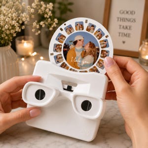 May include: A white plastic stereoscope viewer holding a circular photo reel. The reel features multiple small images and a central larger photograph of two people with the text I love you and a heart symbol printed in white script on the image.
