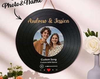 Op de afbeelding: Een zwarte vinyl platen wanddecoratie met een aangepaste foto van een stel, de namen "Andrew & Jessica" in gouden letters, en de tekst "Custom Song" en "Custom Artist Name".