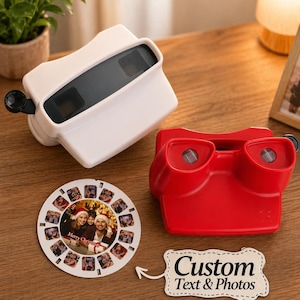 Pode incluir: Dois brinquedos View-Master, um branco e outro vermelho, estão em uma superfície de madeira, acompanhados por um rolo de fotos personalizado. O rolo apresenta uma foto de família de Natal e as palavras "Merry Christmas". Uma placa com "Custom Text & Photos" também está presente.
