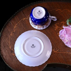 May include: A Royal Albert fine bone china teacup and saucer set. The teacup is cobalt blue with gold filigree and a white handle. The saucer is white with a scalloped edge. The set is on a wooden table with a floral design.