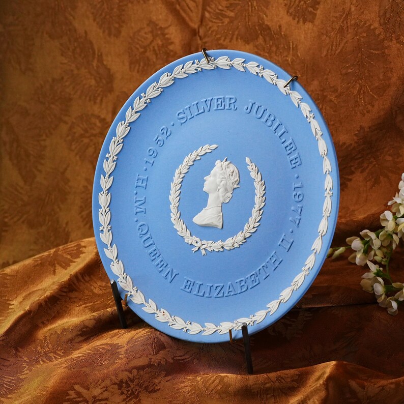 May include: A light blue commemorative plate with white detailing. The plate features a profile of a person within a laurel wreath, surrounded by text that reads "SILVER JUBILEE 1952-1977 QUEEN ELIZABETH II". The plate is displayed on a black stand.