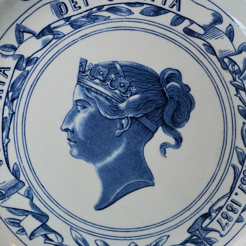 May include: A white and blue ceramic plate featuring a profile portrait of a woman wearing a crown. The portrait is surrounded by a circular border with floral and ribbon designs. The plate has text around the border, including the year 1887.